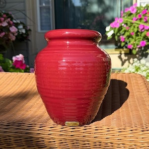 May include: A red ceramic vase with a wide body and a narrow neck. The vase has horizontal ridges and a glossy finish. The vase is sitting on a wicker table. The vase is approximately 8 inches tall.
