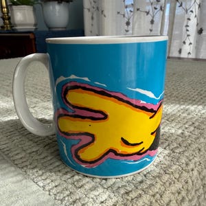 Peut inclure: Une tasse &agrave; caf&eacute; en c&eacute;ramique avec une anse et un bord blancs. La tasse pr&eacute;sente un motif vibrant d'une main jaune avec des contours roses et noirs sur un fond bleu vif. La tasse est pos&eacute;e sur une surface textur&eacute;e.
