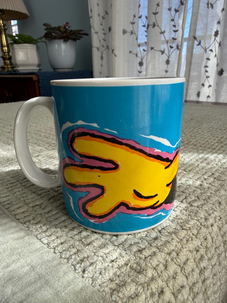 Peut inclure: Une tasse &agrave; caf&eacute; en c&eacute;ramique avec une anse et un bord blancs. La tasse pr&eacute;sente un motif vibrant d'une main jaune avec des contours roses et noirs sur un fond bleu vif. La tasse est pos&eacute;e sur une surface textur&eacute;e.