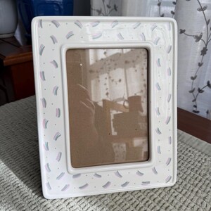 May include: White rectangular picture frame with a decorative pattern of small, colorful brushstrokes in pink, blue, and gray. The frame has a smooth, glossy finish and a brown interior.
