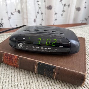 May include: A black GE digital alarm clock displaying 3:02 AM, resting on a vintage brown book. The clock has green LED numbers and AM/PM indicators. The book has worn edges and a textured cover.