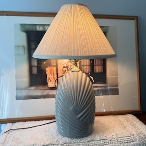 May include: A table lamp featuring a glossy, light gray ceramic base with a textured, fan-like shell pattern. The lamp is topped with a conical, off-white fabric lampshade with fine vertical pleats and a gold-toned metal neck fixture.