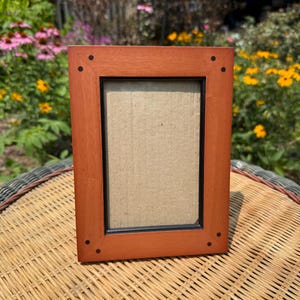 May include: A rectangular wooden picture frame with a warm reddish-brown finish. The frame features a thin black inner border and four small dark circular accents located at each corner of the frame's face. The center contains a plain beige insert.