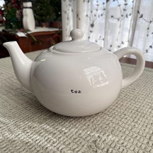 May include: A white ceramic teapot with a lid and handle. The word "tea" is printed in black on the side of the teapot. The teapot is sitting on a textured, light-colored surface.
