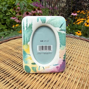 May include: A rectangular picture frame with rounded corners featuring a watercolor-style pattern in shades of green, yellow, and purple. The center has an oval opening with a light blue insert printed with the text SIZE: 2" x 3" and a barcode.