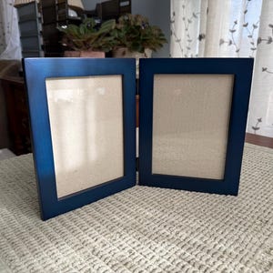 May include: A navy blue, two-panel picture frame. Each panel has a rectangular opening with a light beige backing. The frame is made of wood and is displayed open on a textured, off-white surface.