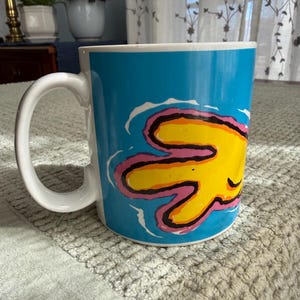 Peut inclure: Une tasse en c&eacute;ramique blanche avec une grande anse incurv&eacute;e. La tasse pr&eacute;sente un fond bleu vif avec un motif abstrait jaune et rose. La tasse est pos&eacute;e sur une surface textur&eacute;e.