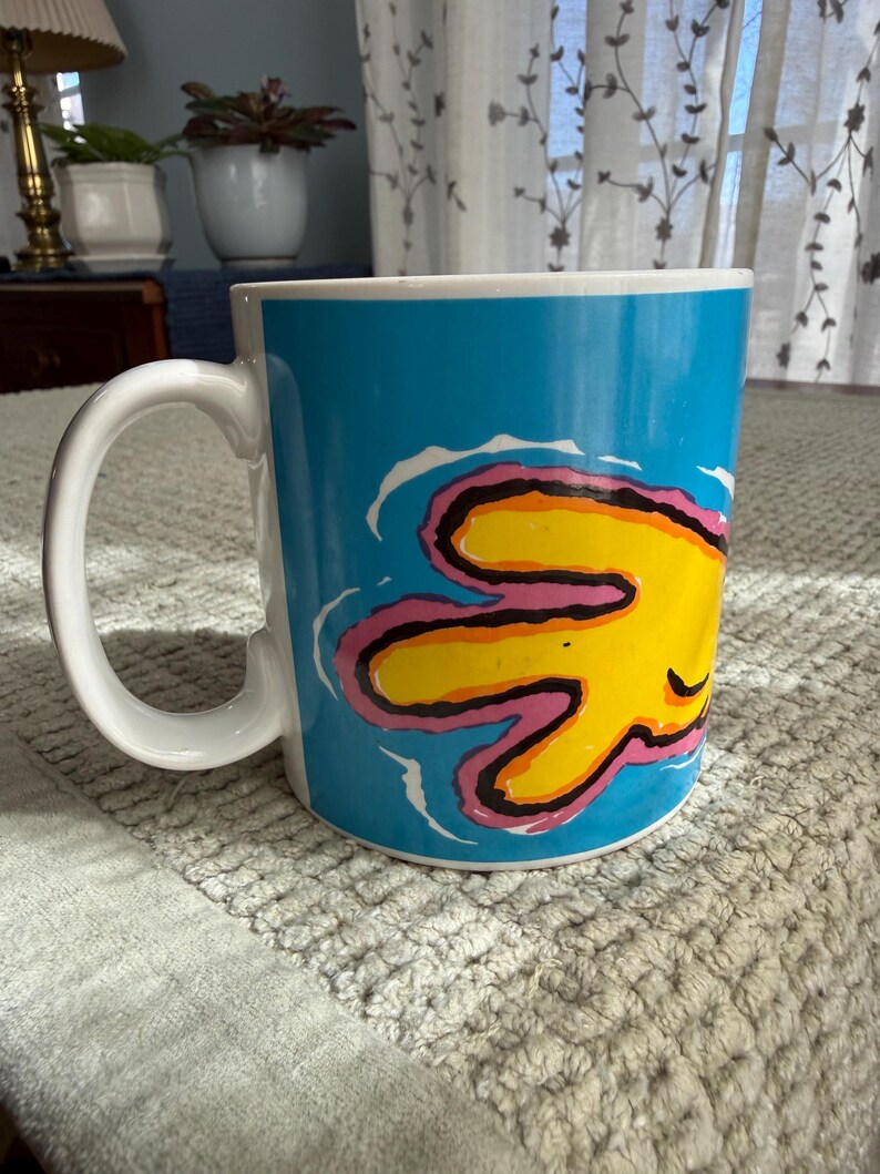 Peut inclure: Une tasse en c&eacute;ramique blanche avec une grande anse incurv&eacute;e. La tasse pr&eacute;sente un fond bleu vif avec un motif abstrait jaune et rose. La tasse est pos&eacute;e sur une surface textur&eacute;e.