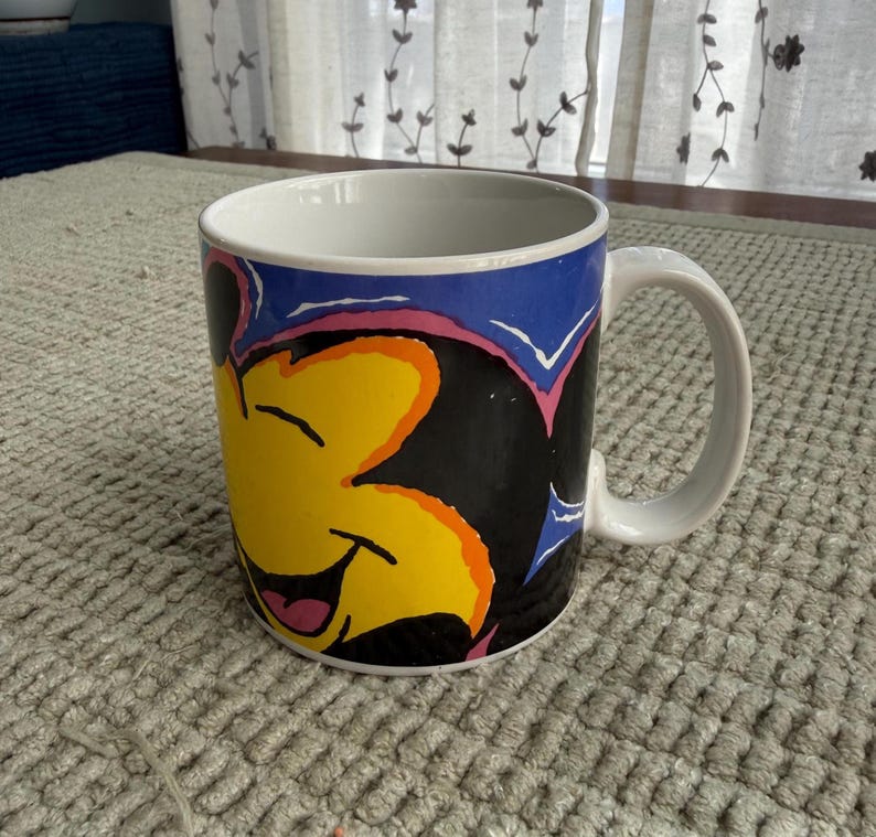 Peut inclure: Mug en c&eacute;ramique blanche avec un personnage de dessin anim&eacute; souriant aux accents jaunes, noirs, roses et bleus. La tasse a une anse blanche et un bord bleu. Le design est une illustration color&eacute;e.