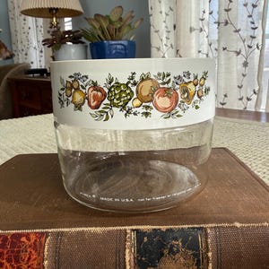 May include: A clear glass Pyrex jar with a white band featuring a colorful fruit and vegetable design. The jar is sitting on a brown book. The text "MADE IN U.S.A." is visible on the jar.