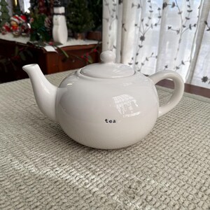 May include: A white ceramic teapot with a rounded body, a curved spout, and a loop handle. The word "tea" is printed in black on the front. The teapot has a lid with a small knob. The teapot is on a textured, light-coloured surface.