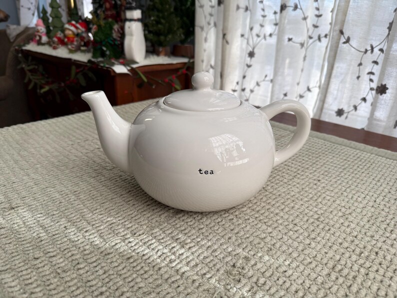 May include: A white ceramic teapot with a rounded body, a curved spout, and a loop handle. The word "tea" is printed in black on the front. The teapot has a lid with a small knob. The teapot is on a textured, light-coloured surface.