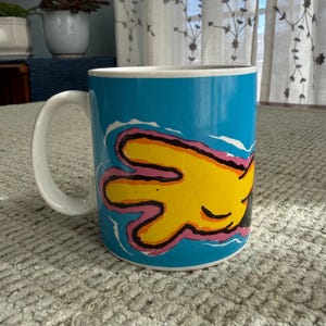Peut inclure: Mug en c&eacute;ramique avec une anse blanche et un ext&eacute;rieur bleu vif. Le mug pr&eacute;sente un motif de main jaune avec des contours roses et noirs. Convient aux boissons chaudes ou froides. Capacit&eacute; d'environ 300 ml.