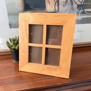 May include: A light-colored wooden picture frame with a grid design. The frame has four openings and a brown backing. The frame is sitting on a dark wooden surface. A small succulent plant in a black pot is to the left of the frame.