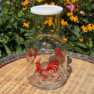 May include: A clear glass pitcher featuring a red rooster graphic printed on the front. The pitcher has a wide base, a narrow neck, and a solid white plastic lid. The glass surface has a subtle textured finish throughout the body of the vessel.
