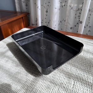 May include: A black rectangular tray with a slightly curved edge and a cut-out on one side. The tray is made of a glossy material and sits on a textured, off-white surface. The tray has the word "TENSI" printed on it.