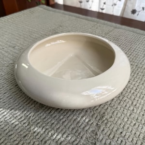May include: A round, off-white ceramic bowl with a wide rim. The bowl has a smooth, glossy finish and a shallow interior. The bowl is sitting on a light gray textured surface. This bowl is suitable for decorative or functional use.