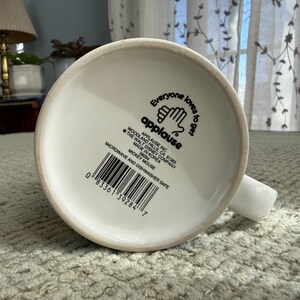 Peut inclure: Mug en c&eacute;ramique blanche avec une anse, portant l'inscription "Everyone loves to get applause" et un code-barres. Le mug est marqu&eacute; comme allant au micro-ondes et au lave-vaisselle. Le fond du mug est visible.