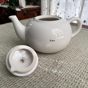 May include: A white ceramic teapot with a matching lid, on a textured surface. The teapot has a rounded body, a curved spout, and a handle. The word "tea" is printed on the side of the teapot.