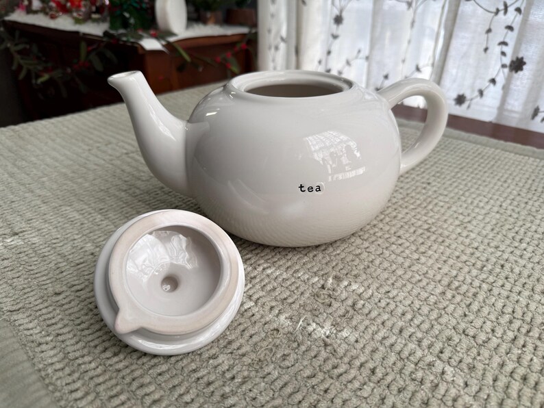 May include: A white ceramic teapot with a matching lid, on a textured surface. The teapot has a rounded body, a curved spout, and a handle. The word "tea" is printed on the side of the teapot.