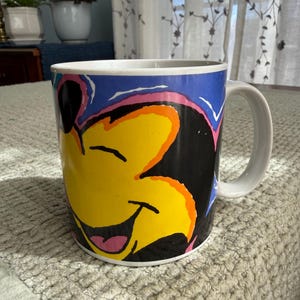 Peut inclure: Mug en c&eacute;ramique blanche avec un visage de dessin anim&eacute; souriant aux accents jaunes, noirs, roses et oranges sur fond bleu. La tasse a une anse blanche et est con&ccedil;ue pour les boissons.