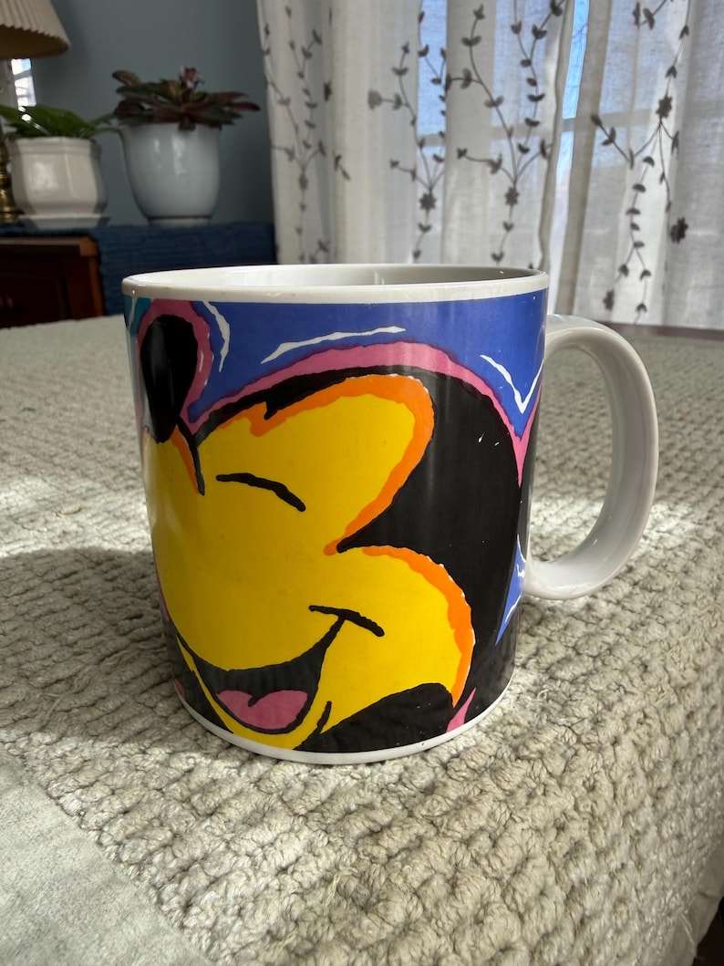 Peut inclure: Mug en c&eacute;ramique blanche avec un visage de dessin anim&eacute; souriant aux accents jaunes, noirs, roses et oranges sur fond bleu. La tasse a une anse blanche et est con&ccedil;ue pour les boissons.