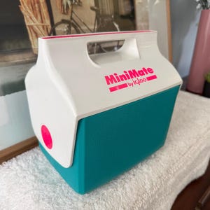 May include: A white and teal MiniMate by Igloo cooler with a pink accent. The cooler has a handle and a pink circular button on the side. The text "MiniMate" is printed in pink.