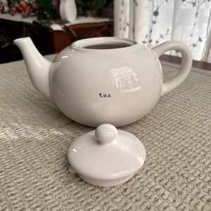 May include: A white ceramic teapot with a matching lid. The teapot has a curved spout and handle. The word "tea" is printed on the side of the teapot. The lid is round with a small knob on top.