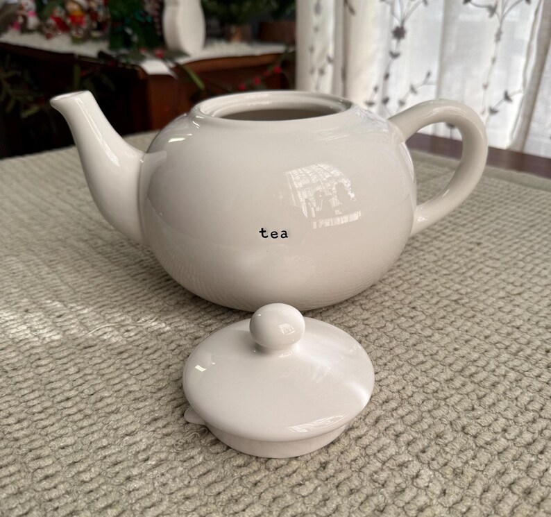 May include: A white ceramic teapot with a matching lid. The teapot has a curved spout and handle. The word "tea" is printed on the side of the teapot. The lid is round with a small knob on top.