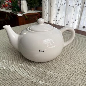 May include: A white ceramic teapot with a rounded body, spout, handle, and lid. The word "tea" is printed in black on the front. The teapot is on a textured, light-coloured surface.