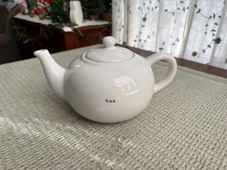 May include: A white ceramic teapot with a rounded body, spout, handle, and lid. The word "tea" is printed in black on the front. The teapot is on a textured, light-coloured surface.