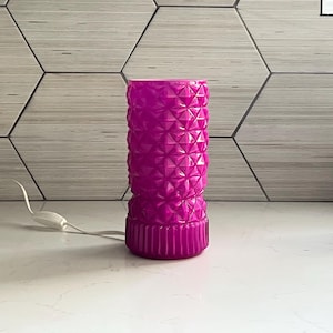 May include: A vibrant magenta table lamp with a textured, geometric pattern. The lamp has a cylindrical shape with a ribbed base and a white power cord. The lamp is on a white surface with a patterned wall in the background.