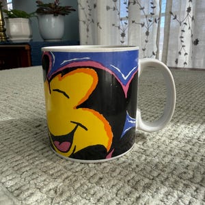 Peut inclure: Tasse en c&eacute;ramique blanche avec un personnage de dessin anim&eacute; souriant, visage jaune, oreilles noires et fond bleu. La tasse a une anse blanche et un design color&eacute;.