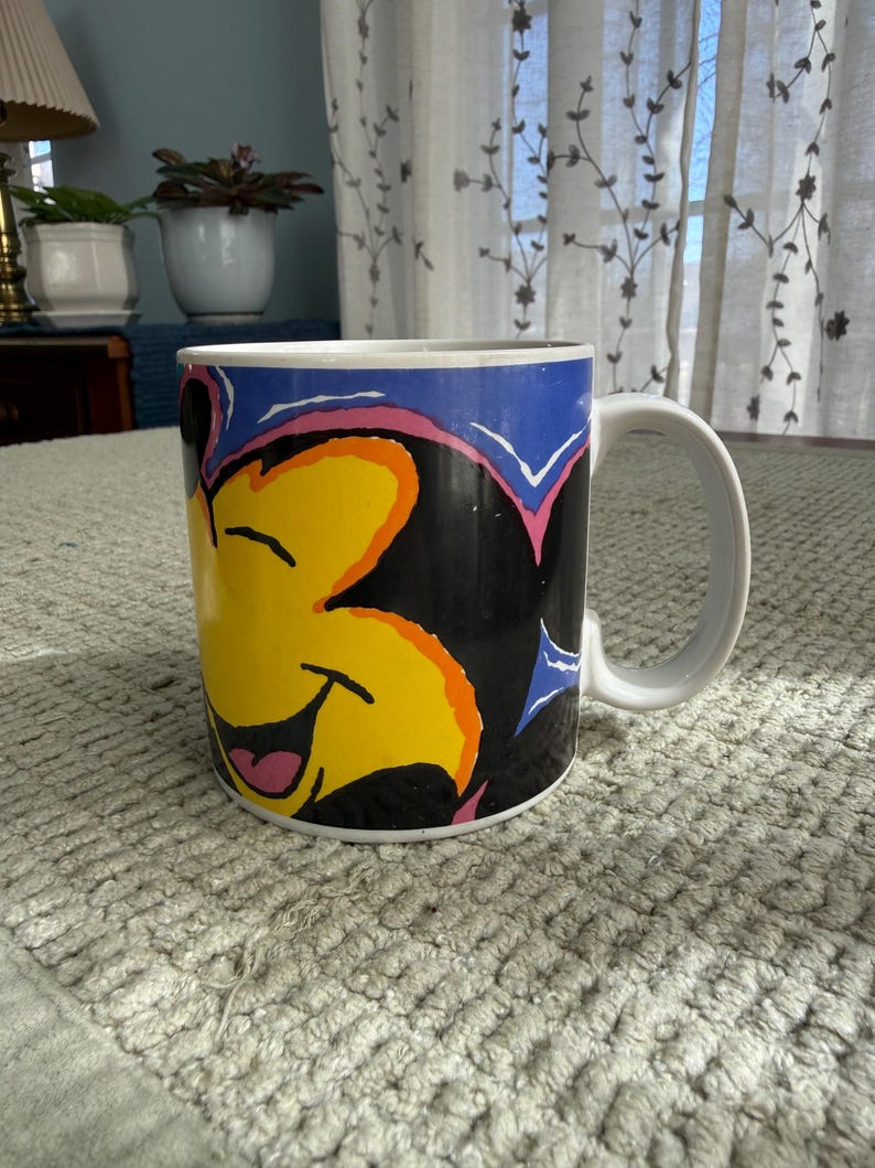 Peut inclure: Tasse en c&eacute;ramique blanche avec un personnage de dessin anim&eacute; souriant, visage jaune, oreilles noires et fond bleu. La tasse a une anse blanche et un design color&eacute;.