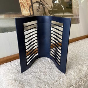 May include: A navy blue, curved photo frame with a series of wave-shaped cutouts. The frame is standing on a white, fluffy surface, with a framed picture of a bicycle and flowers in the background.