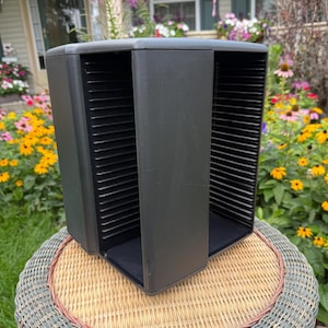 May include: A black plastic media storage rack featuring a vertical design with multiple horizontal slots for organizing discs. The unit has a solid central pillar and open sides, providing a structured frame for holding individual items in a stack.
