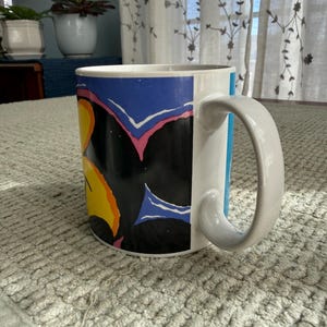 Peut inclure: Mug en c&eacute;ramique blanche avec une anse incurv&eacute;e. Le mug pr&eacute;sente un motif color&eacute; avec des formes noires, bleues, jaunes et roses. Le mug est pos&eacute; sur une surface textur&eacute;e gris clair.