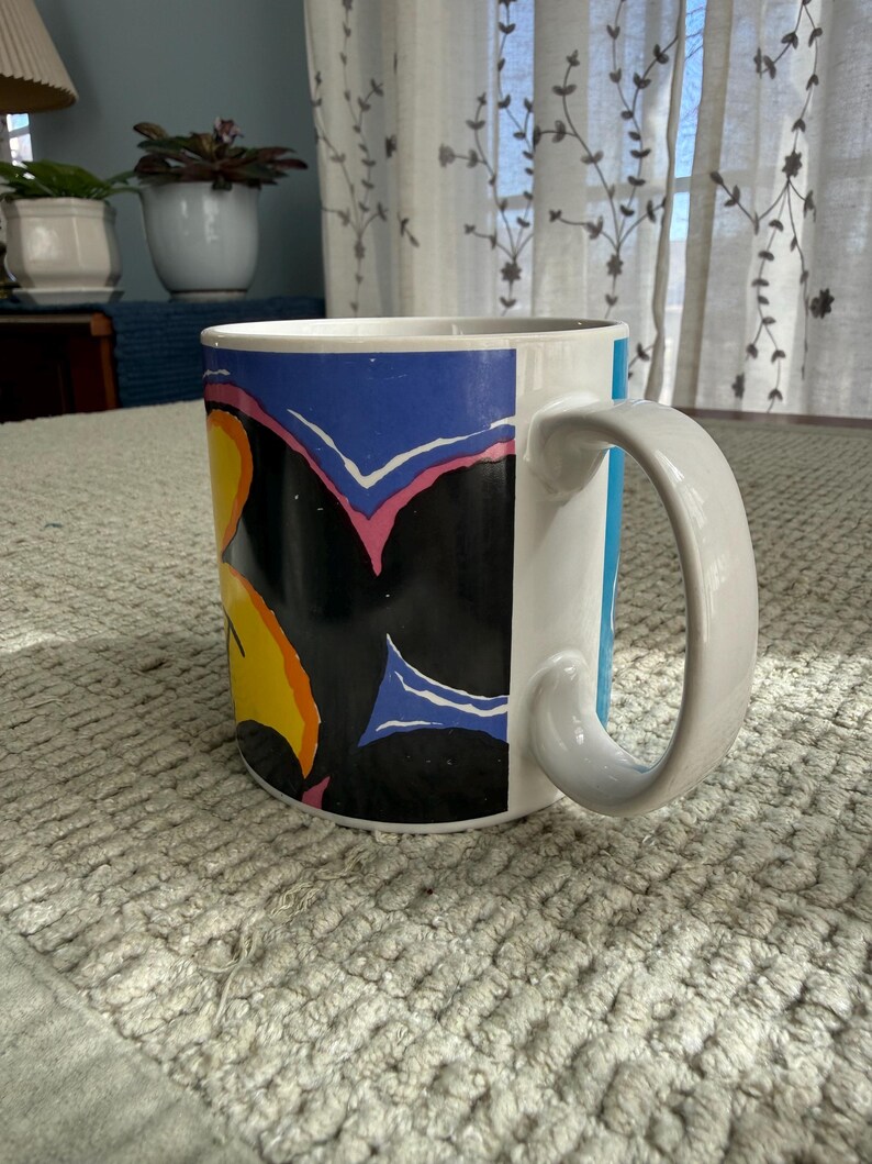 Peut inclure: Mug en c&eacute;ramique blanche avec une anse incurv&eacute;e. Le mug pr&eacute;sente un motif color&eacute; avec des formes noires, bleues, jaunes et roses. Le mug est pos&eacute; sur une surface textur&eacute;e gris clair.