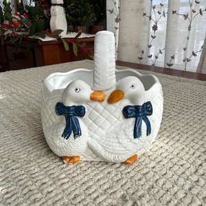 May include: White ceramic basket with two duck figures on the front. The ducks have orange beaks and feet, and dark blue bows. The basket has a handle and a textured, quilted pattern.