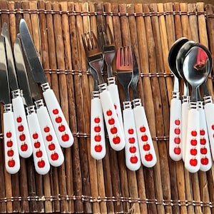 May include: A set of stainless steel cutlery with white handles decorated with red apple designs. The set includes knives, forks, and spoons, arranged on a brown bamboo placemat. The cutlery is designed for dining.