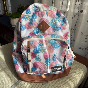May include: White backpack with a repeating pattern of pink and blue pineapples. The backpack has a brown leather bottom and a brown leather patch on the front. The word "trans" is printed on the front pocket.