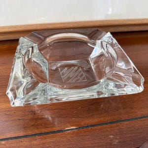 May include: Clear glass ashtray, square with beveled edges. It features a central recessed area and an etched design. The ashtray is made of clear glass and rests on a wooden surface.