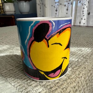Peut inclure: Tasse &agrave; caf&eacute; cylindrique avec un personnage de dessin anim&eacute; souriant. Le personnage a un visage jaune, des oreilles noires et une langue rose. La tasse a un fond bleu avec des accents roses et oranges. La tasse est sur une surface textur&eacute;e grise claire.