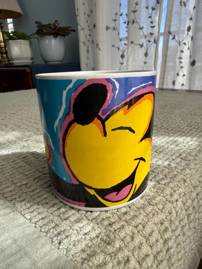 Peut inclure: Tasse &agrave; caf&eacute; cylindrique avec un personnage de dessin anim&eacute; souriant. Le personnage a un visage jaune, des oreilles noires et une langue rose. La tasse a un fond bleu avec des accents roses et oranges. La tasse est sur une surface textur&eacute;e grise claire.