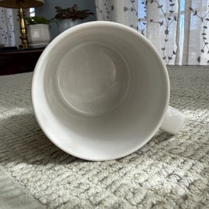 Peut inclure: Une tasse en c&eacute;ramique blanche avec une anse, vue d'en haut. L'int&eacute;rieur de la tasse est visible, montrant une surface lisse et propre. La tasse est pos&eacute;e sur une surface textur&eacute;e, gris clair.