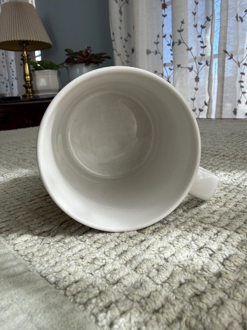 Peut inclure: Une tasse en c&eacute;ramique blanche avec une anse, vue d'en haut. L'int&eacute;rieur de la tasse est visible, montrant une surface lisse et propre. La tasse est pos&eacute;e sur une surface textur&eacute;e, gris clair.