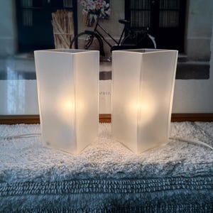 May include: A pair of rectangular table lamps with frosted white glass shades that emit a warm, soft glow. Each lamp features a clean, vertical design with a white power cord extending from the base, providing a steady light source for any space.