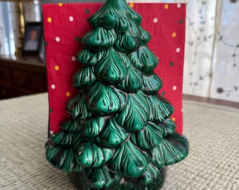 May include: A ceramic Christmas tree napkin holder in glossy emerald green. The tree-shaped holder has detailed leaf textures and holds a red napkin with white and yellow polka dots. A festive holiday decoration.