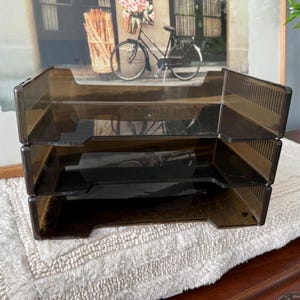 May include: A three-tier desk organizer made of translucent dark brown plastic. The unit features three stacked open trays with ribbed side panels and a cutout front design for easy access to documents, papers, or office supplies.