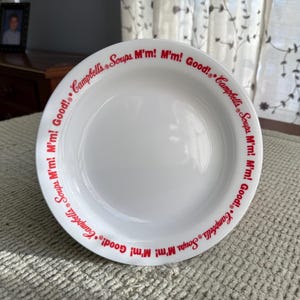 May include: White ceramic soup bowl with a red rim featuring the text "Campbell's Soups M'm! M'm! Good!". The bowl has a wide, shallow design, suitable for serving soup or other dishes. The text is in a classic, retro font.